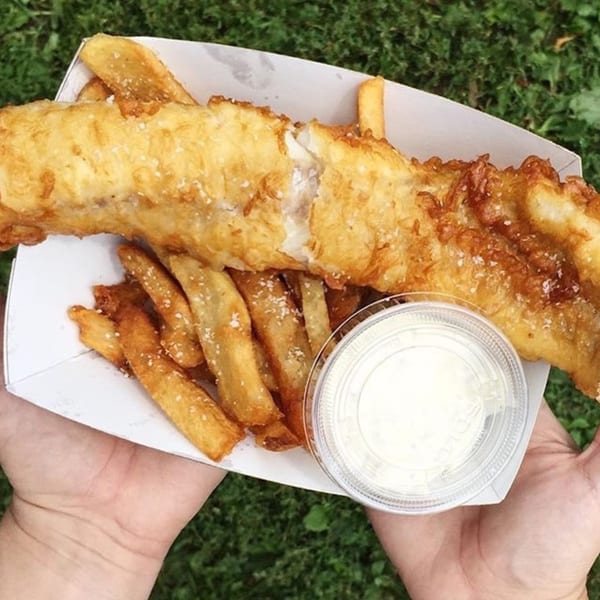 Hands holding a paper tray of golden-brown fish and chips with a side of tartar sauce.