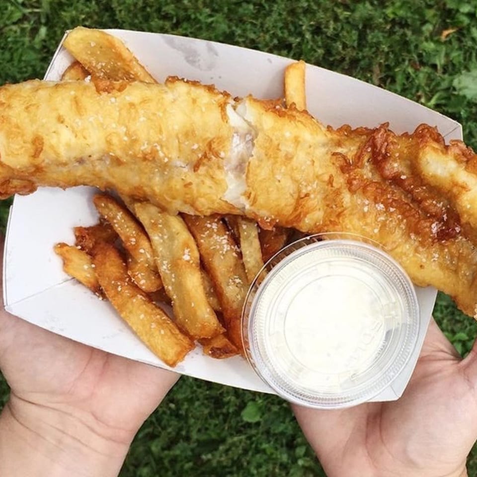 Hands holding a paper tray of golden-brown fish and chips with a side of tartar sauce.