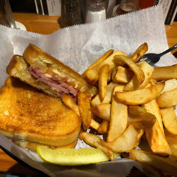 A grilled sandwich with meat and cheese, served with a side of thick-cut fries and a pickle spear.