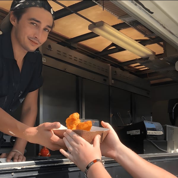 A smiling man in a food truck hands a tray of golden-brown fish and chips to a customer.