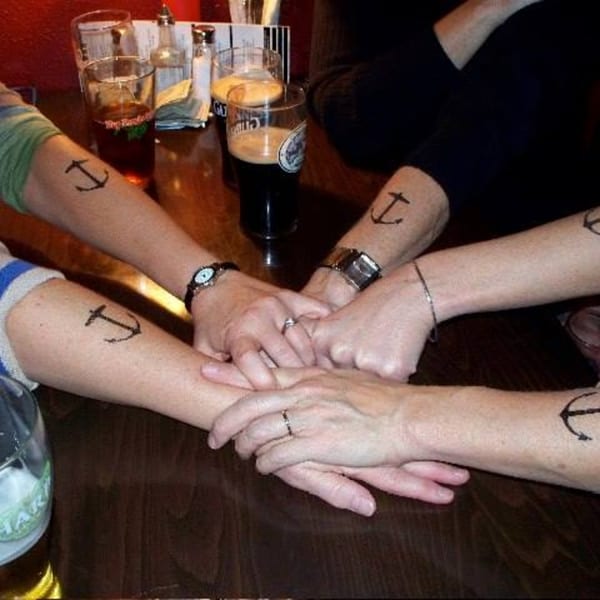 Four people's forearms with anchor tattoos, hands clasped together on a wooden table with drinks.