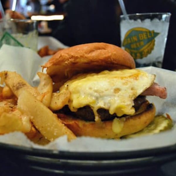 A burger with melted cheese, bacon, and a fried egg, served with fries and a drink.