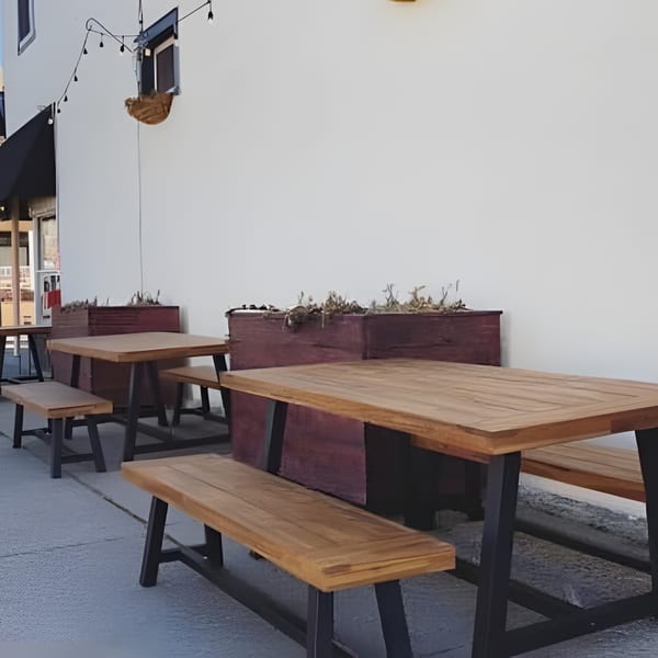 Outdoor patio with wooden picnic tables and benches against a white building with string lights.