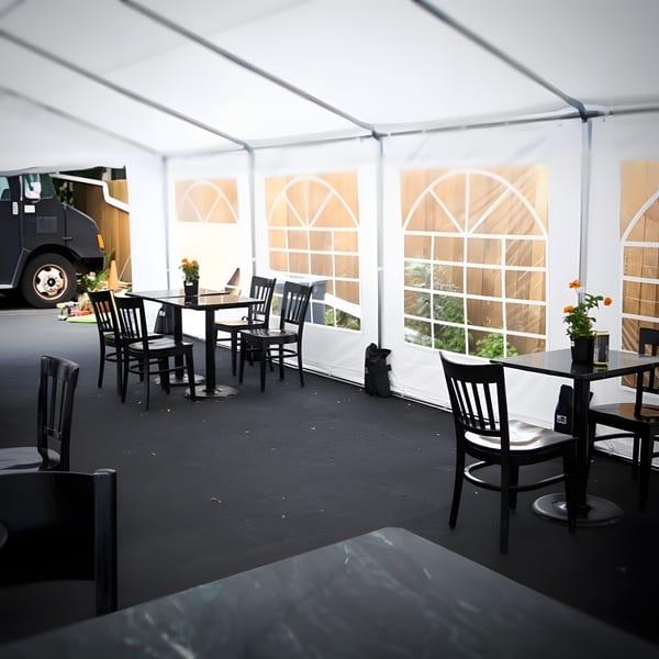 Outdoor dining area under a white tent with black tables and chairs, and a food truck outside.