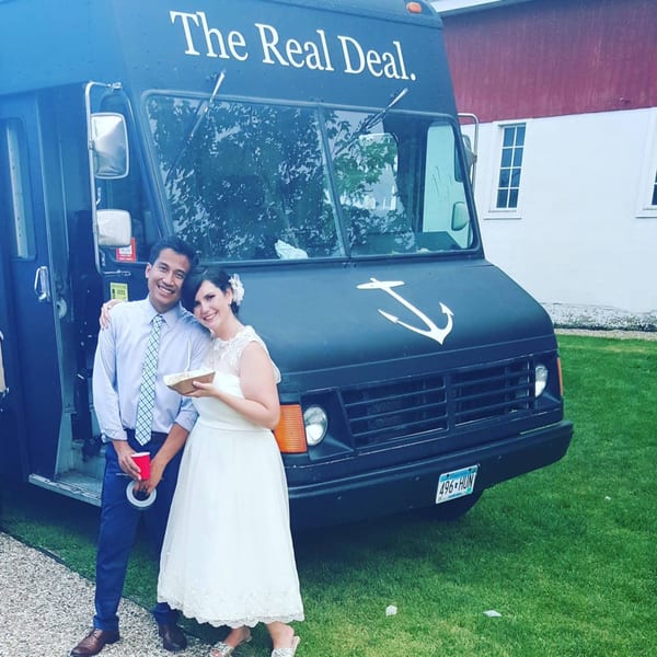 A couple poses in front of The Anchor Fish & Chips food truck, holding food and drinks.